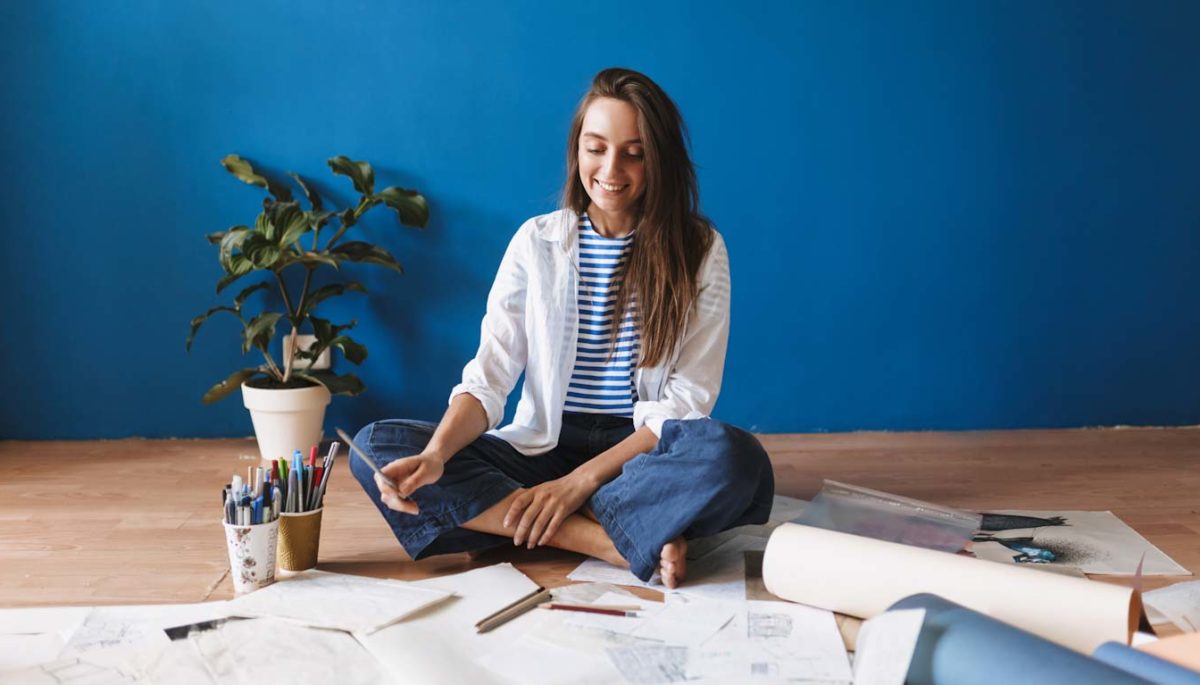 Smiling painter sitting on floor with drawings near over blue ba Image description